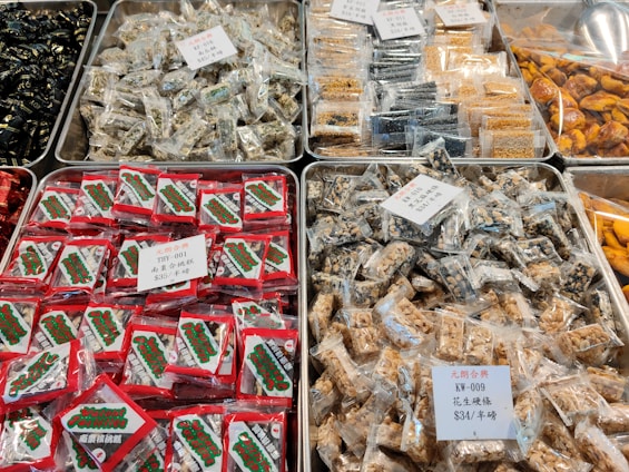 Trays filled with various packaged snacks, each labeled with tags indicating different types and prices. The packages are mostly clear, revealing a variety of candy and nut bars. Some packages are predominantly red with a brand label visible.