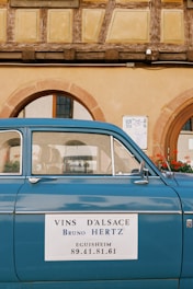 A vintage blue car is parked in front of an old building with traditional timber framing. The car has a sign on the door that reads, 'Vins d'Alsace Bruno Hertz Eguisheim' with a phone number. The building features arched windows and a facade in warm yellow tones.