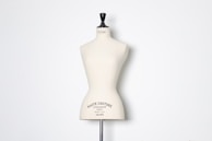 A beige dress form or mannequin torso is placed in front of a plain white background. The top of the mannequin has a black neck and features the inscription 'HAUTE COUTURE STOCKMAN PARIS'. The focus is on the elegant shape and neutral tone of the form.