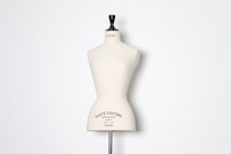 A beige dress form or mannequin torso is placed in front of a plain white background. The top of the mannequin has a black neck and features the inscription 'HAUTE COUTURE STOCKMAN PARIS'. The focus is on the elegant shape and neutral tone of the form.