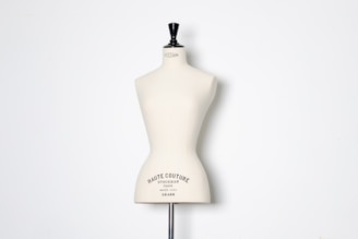 A beige dress form or mannequin torso is placed in front of a plain white background. The top of the mannequin has a black neck and features the inscription 'HAUTE COUTURE STOCKMAN PARIS'. The focus is on the elegant shape and neutral tone of the form.
