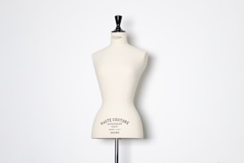 A beige dress form or mannequin torso is placed in front of a plain white background. The top of the mannequin has a black neck and features the inscription 'HAUTE COUTURE STOCKMAN PARIS'. The focus is on the elegant shape and neutral tone of the form.