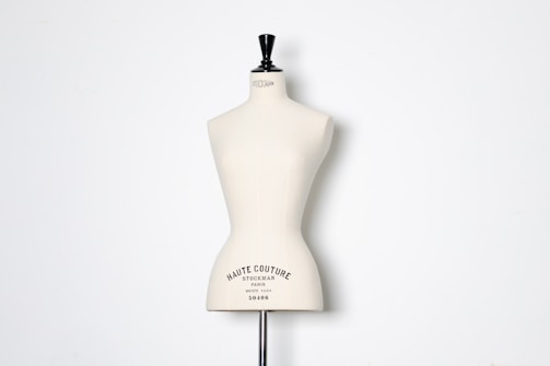 A beige dress form or mannequin torso is placed in front of a plain white background. The top of the mannequin has a black neck and features the inscription 'HAUTE COUTURE STOCKMAN PARIS'. The focus is on the elegant shape and neutral tone of the form.