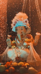 A decorated statue of the Hindu deity Ganesha, adorned with flowers, jewelry, and a traditional headdress. The background features shimmering lights, adding a festive and sacred atmosphere. Fruit offerings and more flowers are placed at the base of the statue.
