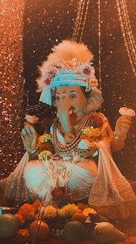 A decorated statue of the Hindu deity Ganesha, adorned with flowers, jewelry, and a traditional headdress. The background features shimmering lights, adding a festive and sacred atmosphere. Fruit offerings and more flowers are placed at the base of the statue.