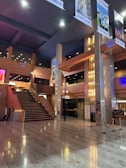 An elegant, spacious indoor area featuring a grand staircase with red carpeting. The space is adorned with modern architectural elements, including tall pillars and sophisticated lighting fixtures. Posters and signs are displayed on the walls, and the floor is composed of polished stone tiles. Ceiling banners display various images and event information.