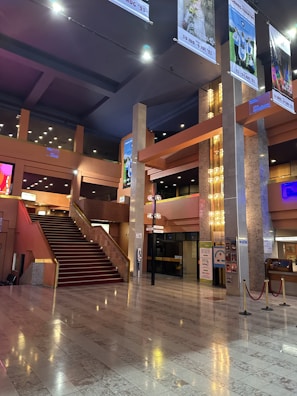 An elegant, spacious indoor area featuring a grand staircase with red carpeting. The space is adorned with modern architectural elements, including tall pillars and sophisticated lighting fixtures. Posters and signs are displayed on the walls, and the floor is composed of polished stone tiles. Ceiling banners display various images and event information.