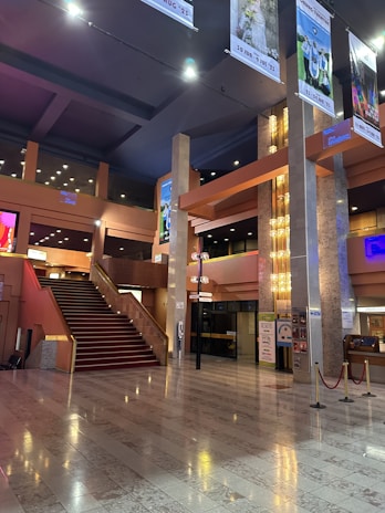 An elegant, spacious indoor area featuring a grand staircase with red carpeting. The space is adorned with modern architectural elements, including tall pillars and sophisticated lighting fixtures. Posters and signs are displayed on the walls, and the floor is composed of polished stone tiles. Ceiling banners display various images and event information.