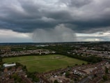 Dark storm clouds overhead with rain visible in the distance over a vast landscape. Below, a residential area with houses and a large green park surrounded by trees is seen.