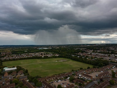 Dark storm clouds overhead with rain visible in the distance over a vast landscape. Below, a residential area with houses and a large green park surrounded by trees is seen.