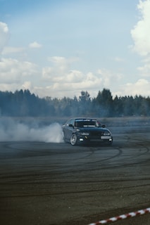 A car is drifting on a track producing plumes of white smoke from its tires. The setting includes an overcast sky with scattered clouds and a background of trees. Tire marks are visible on the asphalt, emphasizing the car's movement.