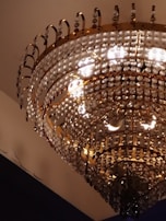 A luxurious chandelier adorned with multiple tiers of sparkling crystal beads and gold accents suspended from a ceiling. The light reflects off the crystals, creating a warm and opulent glow.