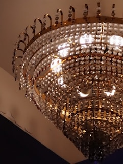 A luxurious chandelier adorned with multiple tiers of sparkling crystal beads and gold accents suspended from a ceiling. The light reflects off the crystals, creating a warm and opulent glow.