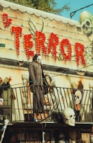A spooky amusement park attraction with a Halloween theme. Bright red lights spell out the word 'TERROR' above the entrance, accompanied by images of skeletons and eerie figures. A mannequin dressed in a dark, hooded cloak and wearing a skull mask stands on a balcony, adding to the haunted ambiance.