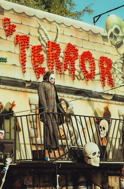 A spooky amusement park attraction with a Halloween theme. Bright red lights spell out the word 'TERROR' above the entrance, accompanied by images of skeletons and eerie figures. A mannequin dressed in a dark, hooded cloak and wearing a skull mask stands on a balcony, adding to the haunted ambiance.