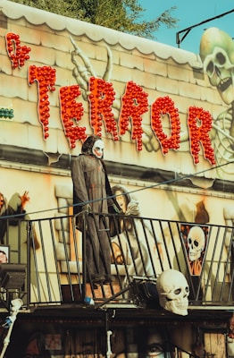 A spooky amusement park attraction with a Halloween theme. Bright red lights spell out the word 'TERROR' above the entrance, accompanied by images of skeletons and eerie figures. A mannequin dressed in a dark, hooded cloak and wearing a skull mask stands on a balcony, adding to the haunted ambiance.