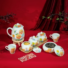 A vibrant tea set featuring a teapot, cups, and containers adorned with colorful floral designs is arranged on a red cloth. Nearby, there are some twigs with red berries and green leaves. The scene also includes a decorative red envelope with floral patterns.
