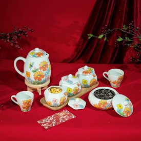 A vibrant tea set featuring a teapot, cups, and containers adorned with colorful floral designs is arranged on a red cloth. Nearby, there are some twigs with red berries and green leaves. The scene also includes a decorative red envelope with floral patterns.