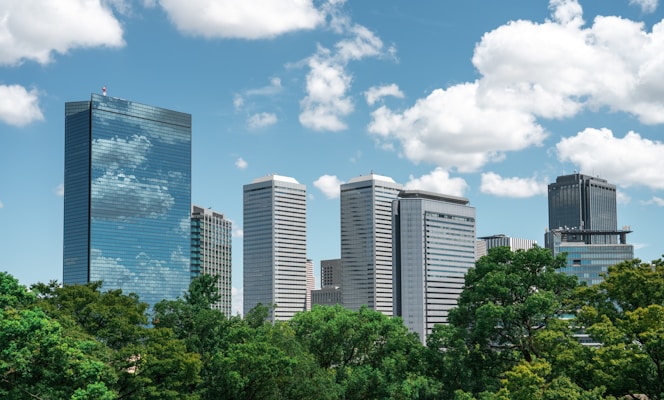 A city skyline with modern skyscrapers set against a bright blue sky filled with scattered white clouds. The buildings have reflective glass facades, and the foreground features lush green trees, suggesting an urban park or green space.