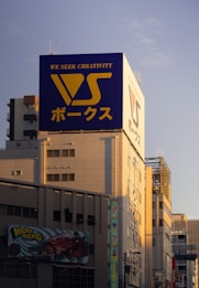 A large building prominently features a billboard with the text 'We Seek Creativity' and Japanese characters, alongside a large logo with the letters 'VS'. The building's facade is bathed in a warm sunset glow, casting shadows across the architecture. An additional advertisement below showcases a drawing of a red car and a cartoon superhero character with the text 'Hero Gang'.
