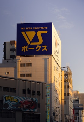 A large building prominently features a billboard with the text 'We Seek Creativity' and Japanese characters, alongside a large logo with the letters 'VS'. The building's facade is bathed in a warm sunset glow, casting shadows across the architecture. An additional advertisement below showcases a drawing of a red car and a cartoon superhero character with the text 'Hero Gang'.