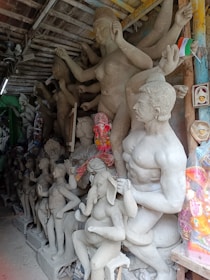 A workshop filled with clay sculptures in various stages of completion. The statues depict human and animal figures, some of which are part of a larger mythological tableau. One sculpture prominently features a figure with an elephant head. The setting appears cluttered with various materials and tools for sculpting. A small Indian flag is visible, suggesting cultural context.