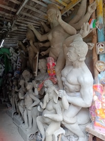 A workshop filled with clay sculptures in various stages of completion. The statues depict human and animal figures, some of which are part of a larger mythological tableau. One sculpture prominently features a figure with an elephant head. The setting appears cluttered with various materials and tools for sculpting. A small Indian flag is visible, suggesting cultural context.
