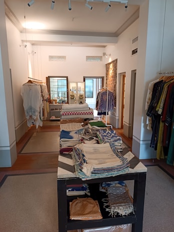 An interior view of a boutique displaying a variety of folded textiles on a central table. Clothes hang on racks along the walls, with a mix of fabrics and patterns. The room features bright lighting, exposed brick accents, and a clean, organized layout.