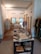 An interior view of a boutique displaying a variety of folded textiles on a central table. Clothes hang on racks along the walls, with a mix of fabrics and patterns. The room features bright lighting, exposed brick accents, and a clean, organized layout.