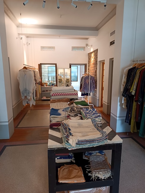 An interior view of a boutique displaying a variety of folded textiles on a central table. Clothes hang on racks along the walls, with a mix of fabrics and patterns. The room features bright lighting, exposed brick accents, and a clean, organized layout.