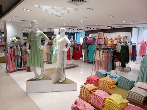 An interior view of a clothing store displaying mannequins dressed in casual outfits. There are racks of colorful clothing in the background and stacks of neatly folded garments in the foreground. The store has a modern and well-lit ambiance.