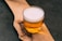 A glass of beer with a frothy white head sits on a wooden surface with a natural edge. The beer is a translucent golden color, and the wood has a rustic, unfinished appearance, with visible grain and a dark knot.