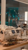 A modern coffee machine sits on a wooden countertop in a well-lit cafe. Two grinders filled with coffee beans are placed beside it. Bright lights and glass windows suggest a clean, contemporary setting.