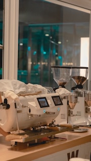 A modern coffee machine sits on a wooden countertop in a well-lit cafe. Two grinders filled with coffee beans are placed beside it. Bright lights and glass windows suggest a clean, contemporary setting.
