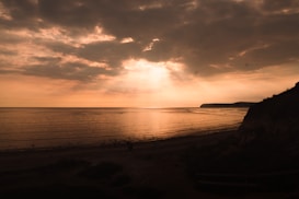 The image captures a serene coastal scene with the sun setting or rising, casting a warm glow across the sky and sea. Dark clouds partially cover the sky, creating dramatic light rays over the calm waters. The shoreline and cliffs are silhouetted against the light, adding depth to the composition.