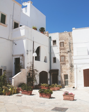 A charming courtyard in a Mediterranean setting, featuring whitewashed buildings with wooden shutters and stone accents. Planters filled with vibrant flowers are arranged on the ground, while climbing plants decorate the walls. The sky is clear and blue, highlighting the warmth and brightness of the scene.