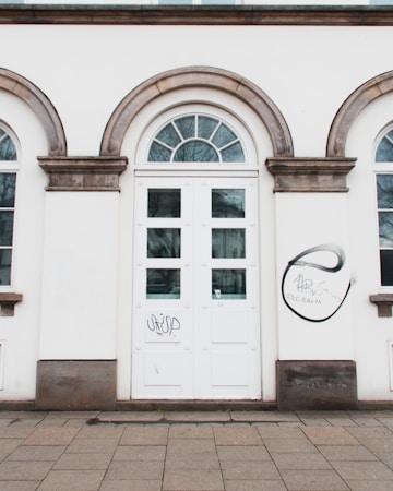 The image features a white building facade with an arched window above a double door. The door is flanked by two narrow windows, and both the door and windows have multiple rectangular panes. Graffiti is present on the wall to the right of the door, including a circular design with some text inside.