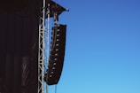 A large speaker system is suspended from a metal framework on the side of a building or stage setup. The sky is clear and blue, providing a stark contrast to the black speakers and metal structure.