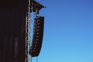 A large speaker system is suspended from a metal framework on the side of a building or stage setup. The sky is clear and blue, providing a stark contrast to the black speakers and metal structure.