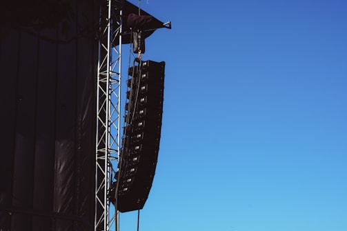 A large speaker system is suspended from a metal framework on the side of a building or stage setup. The sky is clear and blue, providing a stark contrast to the black speakers and metal structure.