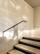 A stairway with steps covered in light-colored tiles leading up, bordered by a wall of white square tiles. Sunlight casts shadows on the wall, creating geometric patterns. A sleek, black handrail runs along the left side, providing a visual guide upwards.
