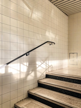 A stairway with steps covered in light-colored tiles leading up, bordered by a wall of white square tiles. Sunlight casts shadows on the wall, creating geometric patterns. A sleek, black handrail runs along the left side, providing a visual guide upwards.