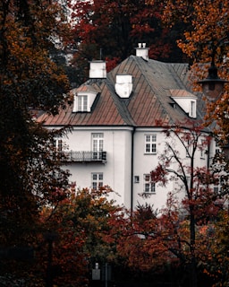 A large, elegant house with a classic architectural style, featuring a muted grey facade and a steep roof with multiple dormer windows. The surrounding trees display vibrant autumn foliage, with shades of red, orange, and yellow. The scene is framed by additional trees, and a streetlamp is partially visible.