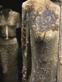 An intricately beaded garment with detailed floral patterns in blue and gold against a shimmering silver background. The focus is on a jacket with rich embroidery, while a similar style top and skirt set are slightly blurred in the background.
