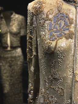 An intricately beaded garment with detailed floral patterns in blue and gold against a shimmering silver background. The focus is on a jacket with rich embroidery, while a similar style top and skirt set are slightly blurred in the background.