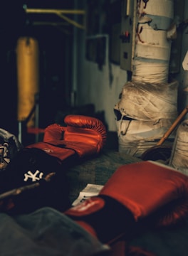 A dimly lit gym or training room featuring a pair of red boxing gloves resting on a surface. The background includes a wrapped pillar and a boxing bag, suggesting an atmosphere of sports training.