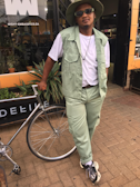 A person dressed in a casual outfit featuring a light green vest and matching pants, paired with a white t-shirt. They are wearing stylish sunglasses and a wide-brimmed hat, leaning against a bicycle with one hand resting on the handlebars. The background has a storefront with glass windows and plants.