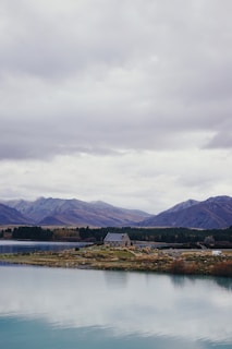A serene lakeside landscape featuring a small stone chapel on the shore, surrounded by grass and trees. The backdrop consists of majestic mountains under a cloudy sky, creating a sense of tranquility and isolation. The water in the foreground is calm and reflects the overcast sky.