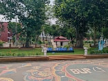 A landscaped outdoor area with large trees and neatly arranged flower beds. A small structure with a red roof sits in the background. There is a decorative design on the paved ground in the foreground, and blue letters spelling out 'ICIT ASD2 23' are displayed prominently.