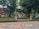 A landscaped outdoor area with large trees and neatly arranged flower beds. A small structure with a red roof sits in the background. There is a decorative design on the paved ground in the foreground, and blue letters spelling out 'ICIT ASD2 23' are displayed prominently.